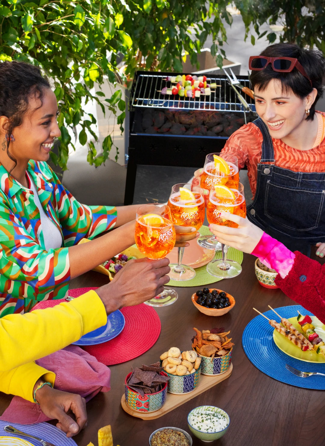 Four friends clinking Aperol Spritz glasses at an outdoor barbecue, colorful outfits and joyful vibe